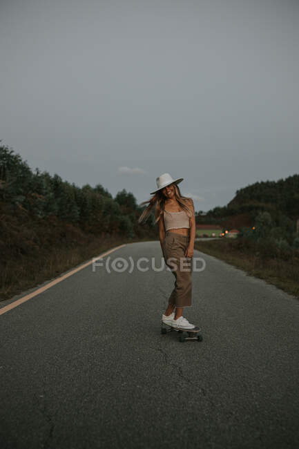 Спортивна жінка в модному одязі верхи на крейсерській дошці вздовж порожньої асфальтової дороги в літній сільській місцевості в сонячний день — стокове фото