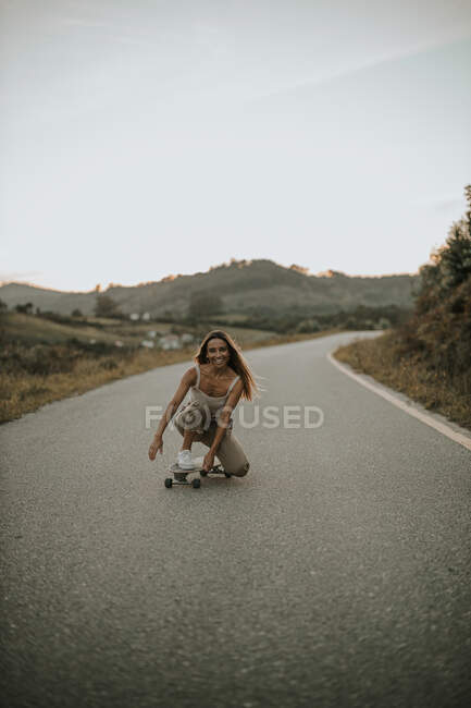 Full body attractive carefree female skater sitting on haunches on skateboard and looking at camera while skating on empty rural road at twilight — Stock Photo