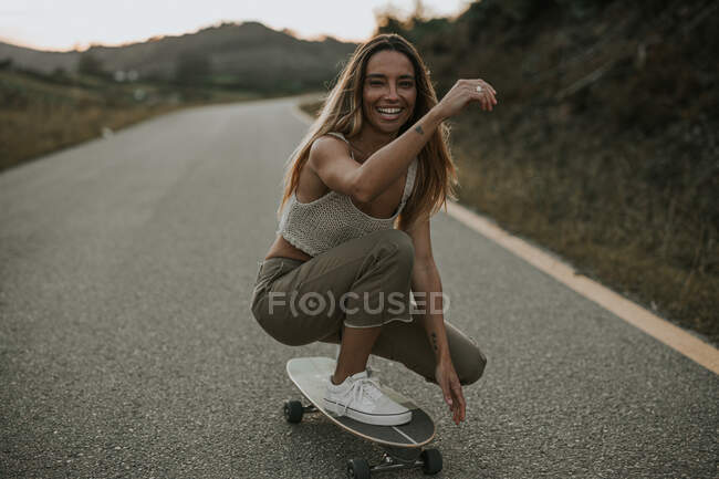 Full body attractive carefree female skater sitting on haunches on skateboard and looking at camera while skating on empty rural road at twilight — Stock Photo