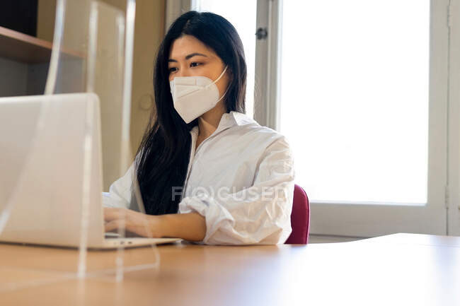 Ejecutivo femenino étnico anónimo en máscara respiratoria escribiendo en netbook en el escritorio en el día soleado - foto de stock