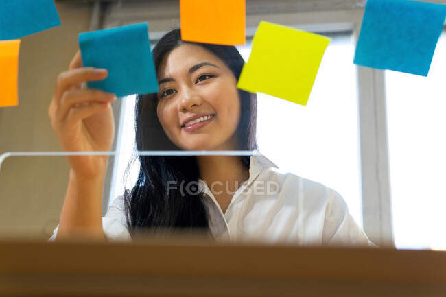 Young glad ethnic female entrepreneur arranging colorful paper stickers on transparent surface in office in daytime — Stock Photo