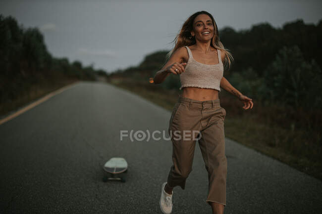 Беззаботная женщина в повседневной одежде бегает по пустой дороге рядом с крейсерской доской в сельской местности в сумерках — стоковое фото