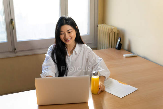 Haut angle de l'entrepreneur ethnique féminin naviguant netbook tout en étant assis avec un document en papier et une bouteille en verre écologique de jus frais — Photo de stock