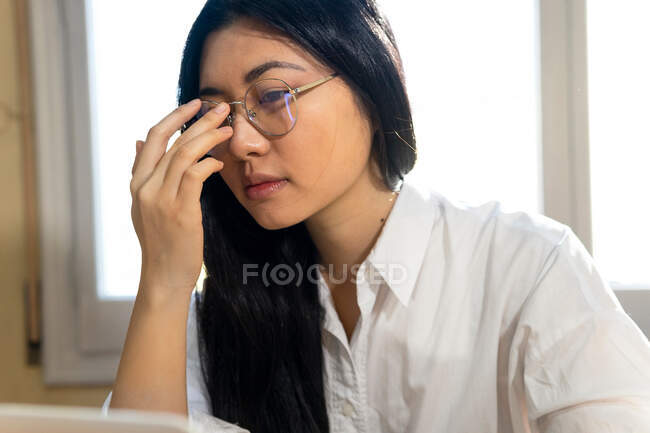 Colheita pensativa étnica feminina autônoma freelancer em óculos trabalhando no gadget em casa — Fotografia de Stock
