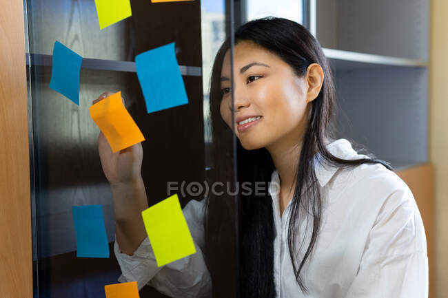 Joven emprendedora étnica alegre organizando pegatinas de papel de colores en la superficie transparente en la oficina durante el día - foto de stock