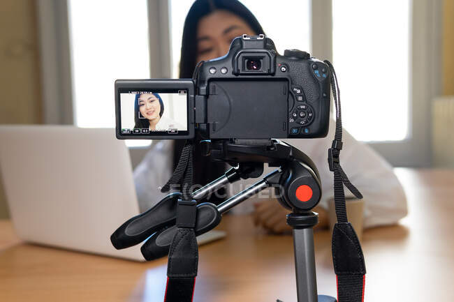 Ritaglia sorridente femmina etnica con netbook registrazione video su fotocamera professionale a tavola in casa — Foto stock