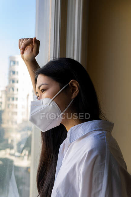 Visão lateral da jovem executiva étnica em máscara respiratória olhando para a janela no espaço de trabalho — Fotografia de Stock