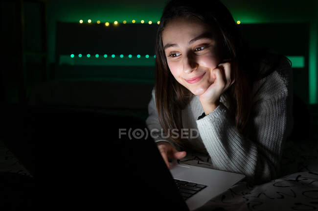 Lächelnder Teenager in lässigem Pullover liegt im Schlafzimmer, während er im dunklen Raum im Netbook blättert — Stockfoto