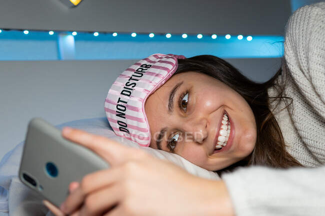 Cheerful dark haired female in sleep mask on head browsing mobile phone while resting on bed and looking at screen — Stock Photo