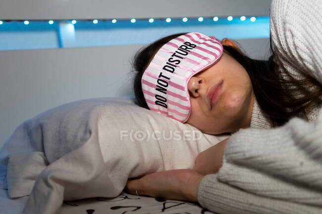 Jovem fêmea de cabelos escuros em máscara de sono deitado no travesseiro no quarto com lâmpadas na cama — Fotografia de Stock