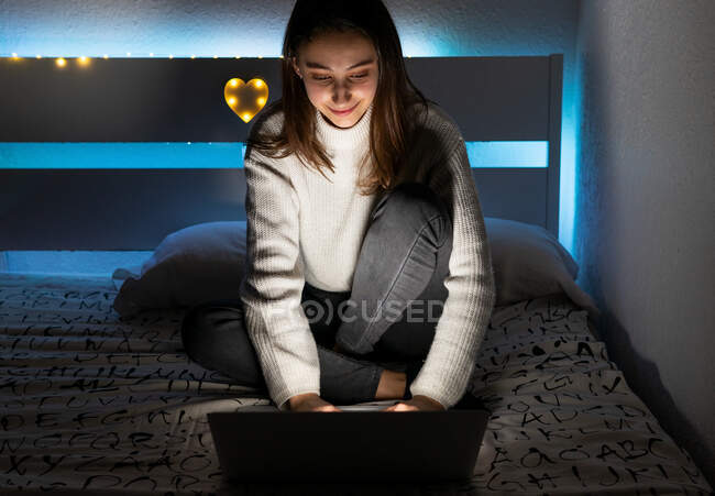 Улыбающийся подросток в обычном свитере сидит на спальне, просматривая нетбук в темной комнате — стоковое фото