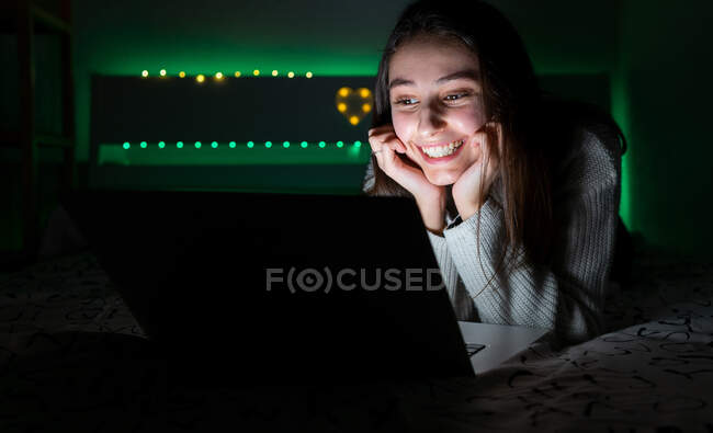Adolescent souriant en pull décontracté couché sur la chambre à coucher tout en naviguant netbook dans la chambre noire — Photo de stock
