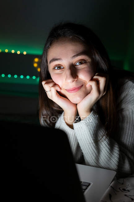 Smiling teenager in casual sweater lying on bedroom while browsing netbook in dark room — Stock Photo