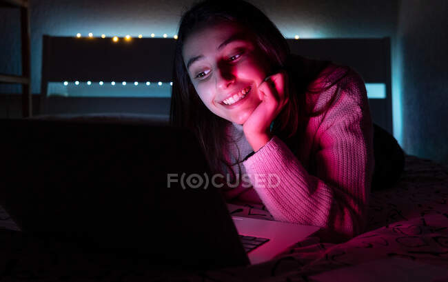 Adolescente sorridente em camisola casual deitado no quarto enquanto navega netbook no quarto escuro — Fotografia de Stock