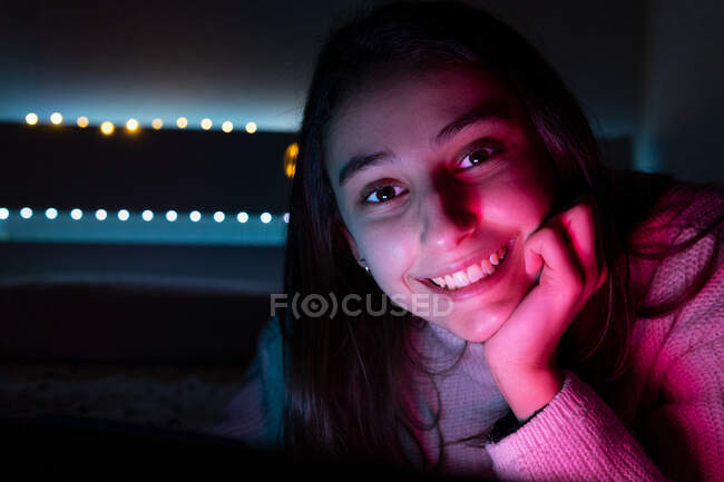 Adolescente de cabelos escuros expressivo em suéter sentado sob luz brilhante e dente sorrindo enquanto olha para a câmera — Fotografia de Stock