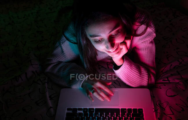 Усміхнений підліток у повсякденному светрі лежить на спальні під час перегляду нетбука в темній кімнаті — стокове фото