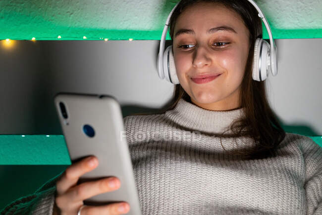 Crop чарівна жінка в светрі слухає музику з навушниками під час перегляду мобільного телефону на ліжку — стокове фото