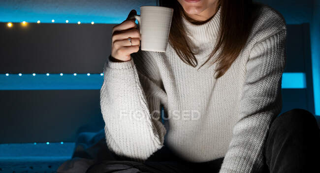 Crop anonymous dark haired female in sweater drinking hot aromatic beverage while resting at home — Stock Photo