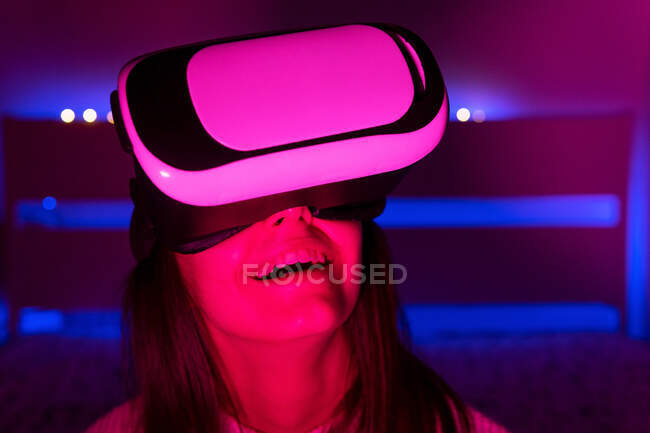 Young dark haired female in VR goggles looking around while sitting in room with vivid illumination — Stock Photo