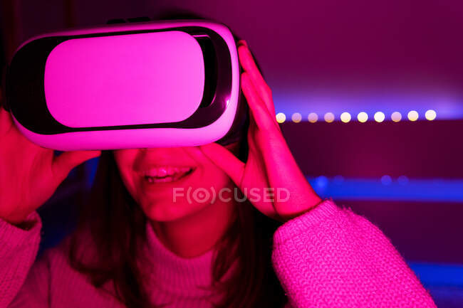Jovem fêmea de cabelos escuros em óculos VR olhando ao redor enquanto sentado na sala com iluminação vívida — Fotografia de Stock