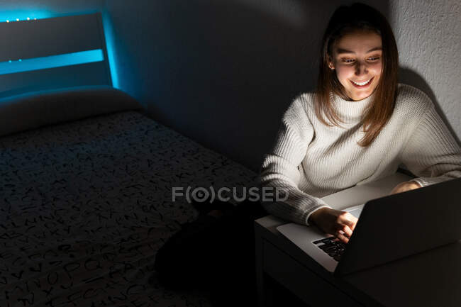 Adolescente sorridente in maglione casual seduto sulla camera da letto durante la navigazione netbook in camera oscura — Foto stock