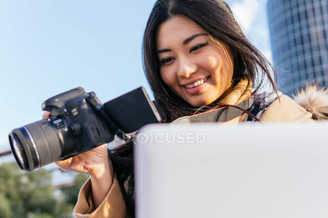 Baixo ângulo de concentrado étnico asiático fotógrafo feminino vestindo outerwear quente navegação netbook enquanto trabalhava remotamente — Fotografia de Stock