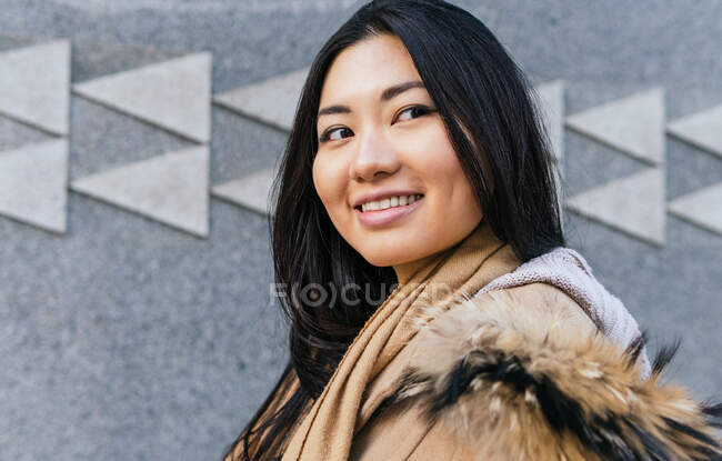 Mulher étnica positiva em outerwear quente em pé contra a parede de azulejos com ornamento geométrico e olhando para longe — Fotografia de Stock
