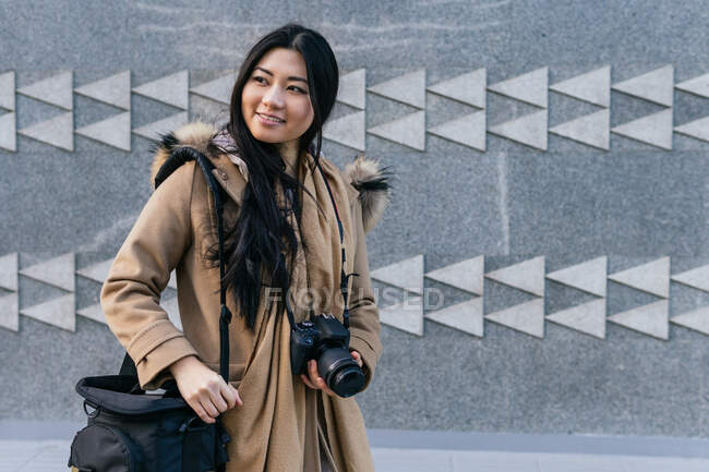 Positive Asian female photographer wearing warm coat with photo camera standing against stone wall with geometric decor — Stock Photo