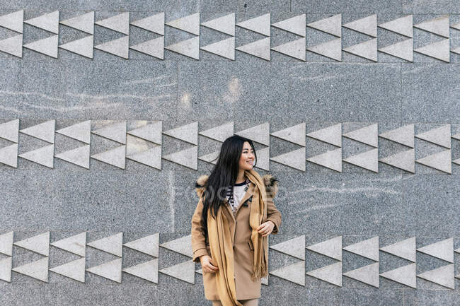 Mulher étnica positiva em outerwear quente em pé contra a parede de azulejos com ornamento geométrico e olhando para longe — Fotografia de Stock