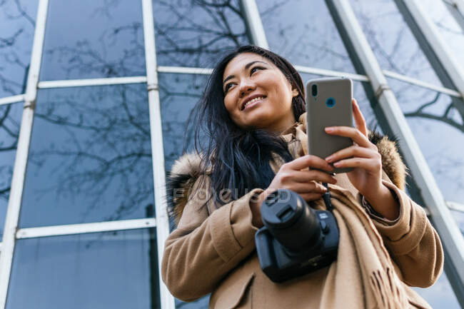 Desde abajo de la sonriente hembra asiática navegando teléfono móvil de pie con cámara de fotos contra la pared de vidrio - foto de stock