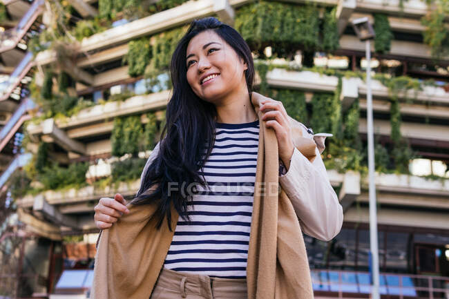 Low angle of smiling ethnic female wearing coat standing against modern building with green plants — Stock Photo