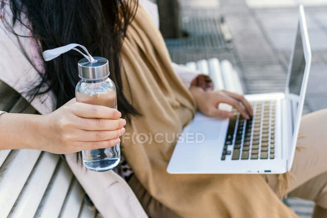 Анонимная женщина, держащая в руках экологически чистую бутылку с водой, работает дистанционно на ноутбуке, сидя на скамейке на улице — стоковое фото