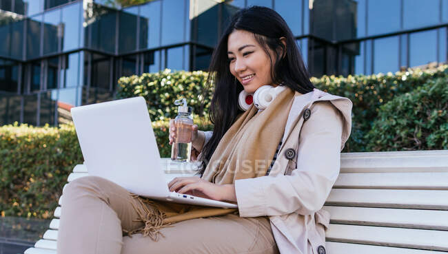 Sonriente hembra asiática en auriculares inalámbricos sentados en el banco con botella de agua ecológica y netbook de navegación mientras trabaja de forma remota - foto de stock