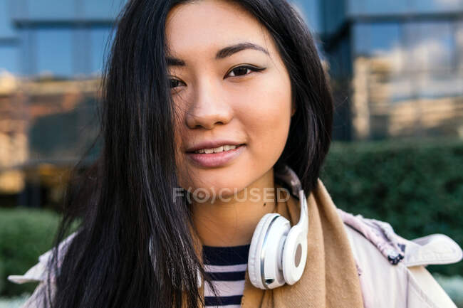 Sonriente mujer étnica usando abrigo con auriculares alrededor del cuello de pie contra el edificio moderno - foto de stock