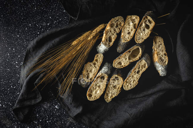 Сверху аппетитный хрустящий хлеб возле пшеничных шипов и темной ткани на столе — стоковое фото
