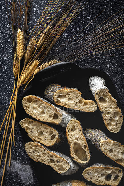 Desde arriba de apetitoso pan crujiente cerca de espigas de trigo y tela oscura en la mesa - foto de stock