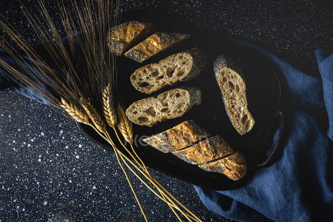 Desde arriba de apetitoso pan crujiente cerca de espigas de trigo y tela oscura en la mesa - foto de stock