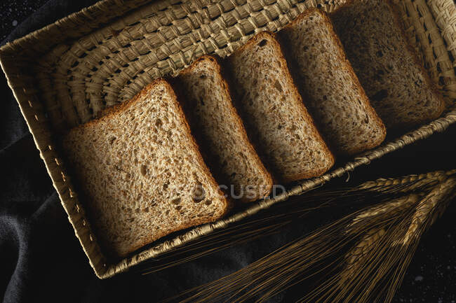 Vista superior de pan de centeno casero fresco cerca de cuchillo en canasta de mimbre y espigas de trigo en la mesa - foto de stock