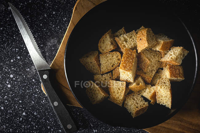 Trozos de pan crujiente en tazón cerca de espigas de trigo en textil negro en la habitación - foto de stock