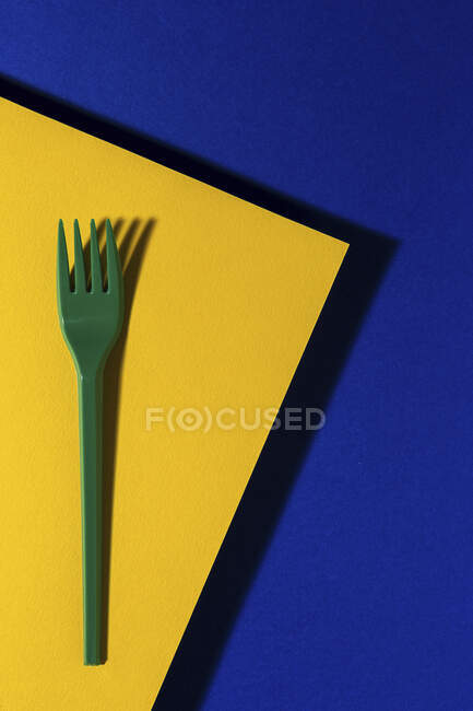Vista aérea de tenedor ecológico verde brillante cerca de la hoja de cartón amarillo sobre fondo azul - foto de stock