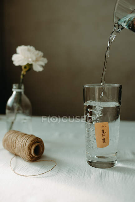 Water pouring into glassware with inscription i miss you placed near skein of thread and blooming carnation — Stock Photo