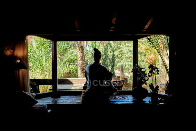 Vue arrière d'un homme anonyme pratiquant le yoga assis dans la maison contre un jardin d'été avec des palmiers — Photo de stock