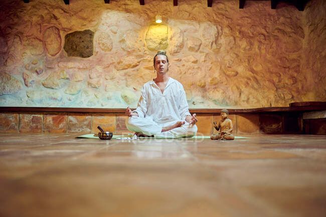 Jovem macho concentrado com olhos fechados sentado na pose Padmasana enquanto pratica ioga perto da estatueta de Buda — Fotografia de Stock