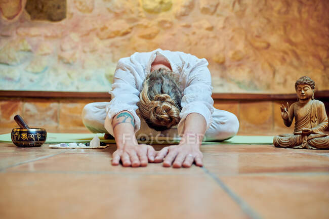 Anonymous flexible male with tattoo practicing yoga between Buddha statuette and bowl gong against stone wall — Stock Photo