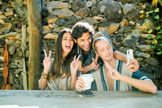 Jeunes heureux partenaires multiethniques prenant autoportrait sur appareil photo à table contre les plantes grimpantes dans la cour — Photo de stock
