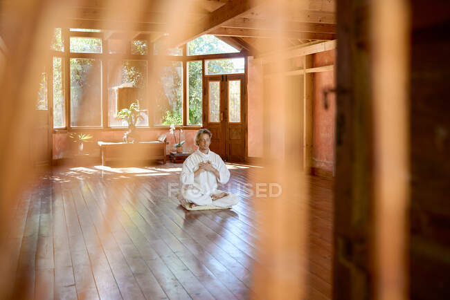 Молодий розумний чоловік босоніж, сидячи в позі лотоса на подушці з закритими очима практикує йогу на підлозі біля миски гонг і статуетки Будди — стокове фото
