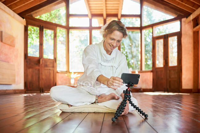 Jeune homme joyeux en vêtements blancs avec téléphone portable assis avec les jambes croisées sur l'oreiller dans le hall de la maison — Photo de stock