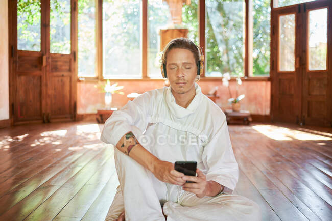Молодий уважний чоловік у білому вбранні з мобільним телефоном сидить на подушці, слухаючи курс йоги із закритими очима від гарнітури — стокове фото