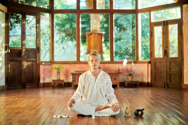 Молодой внимательный мужчина босиком сидит в позе лотоса и практикует йогу на полу рядом со статуэткой Будды и чашечкой. — стоковое фото