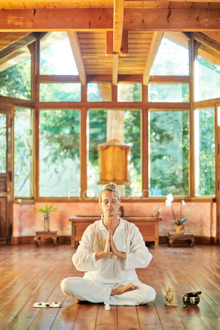 Молодой внимательный мужчина босиком сидит в позе лотоса и практикует йогу на полу рядом со статуэткой Будды и чашечкой. — стоковое фото